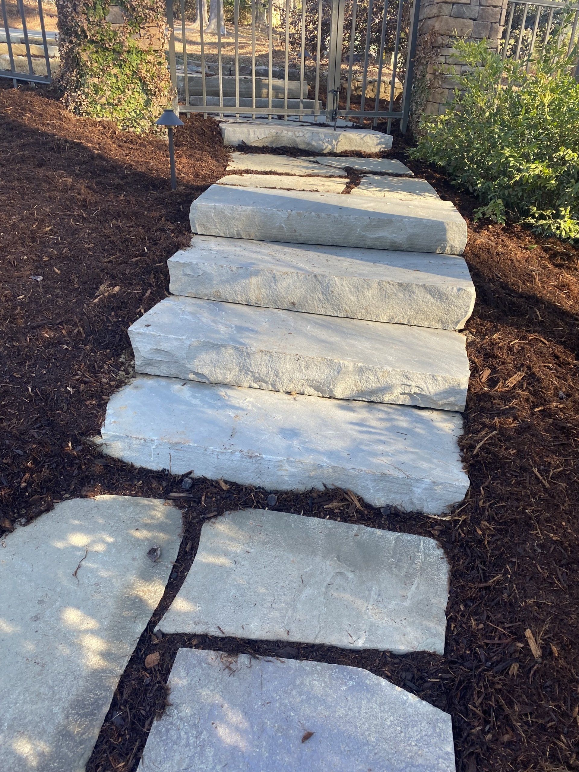 A set of light gray stone stairs leading up to a metal gate, surrounded by dark brown mulch and green foliage.