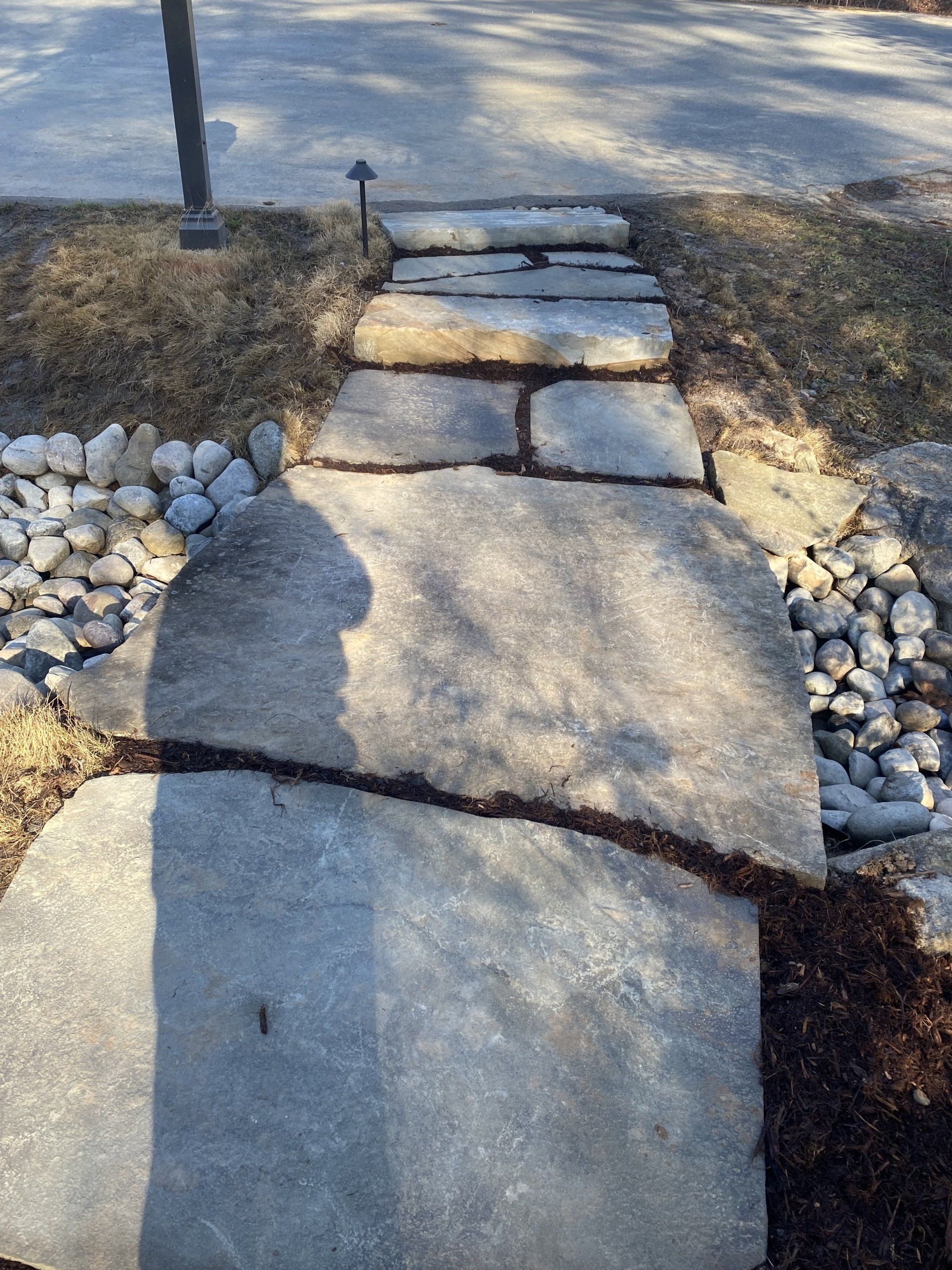 A stone walkway made of large, flat, uneven slabs leading toward a paved road, flanked by decorative river rocks.
