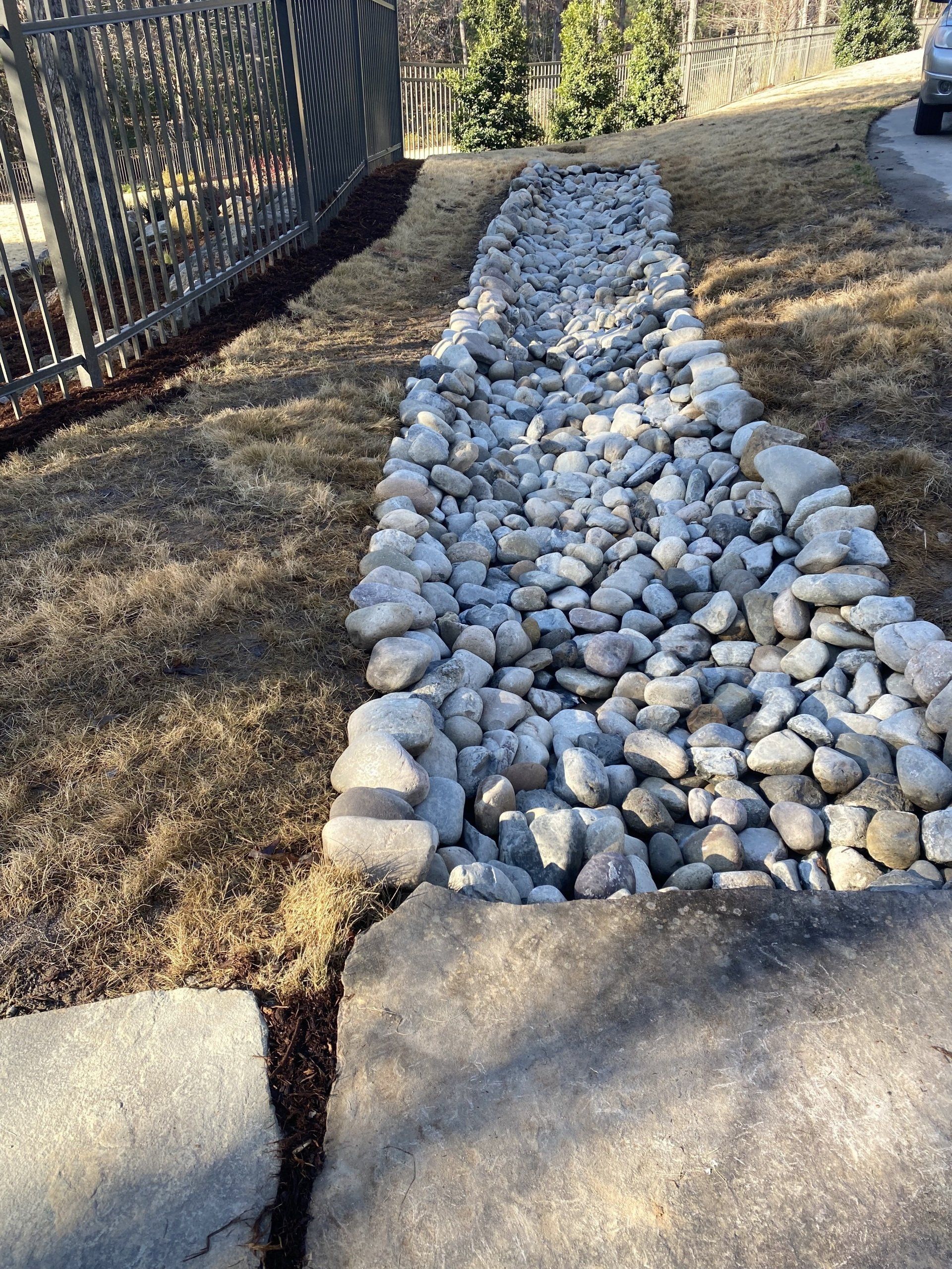 A dry creek bed of light gray river rocks running along a fence line next to a grass lawn and concrete driveway.