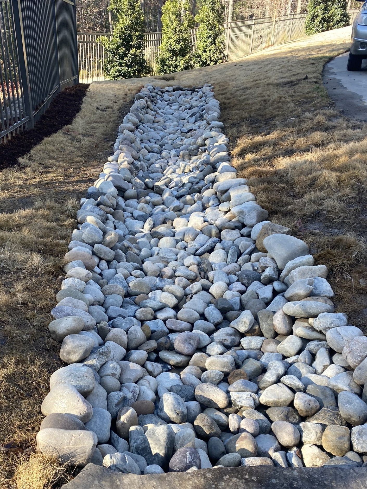 A rock-filled drainage trench runs through a grassy lawn between a metal fence and a driveway.