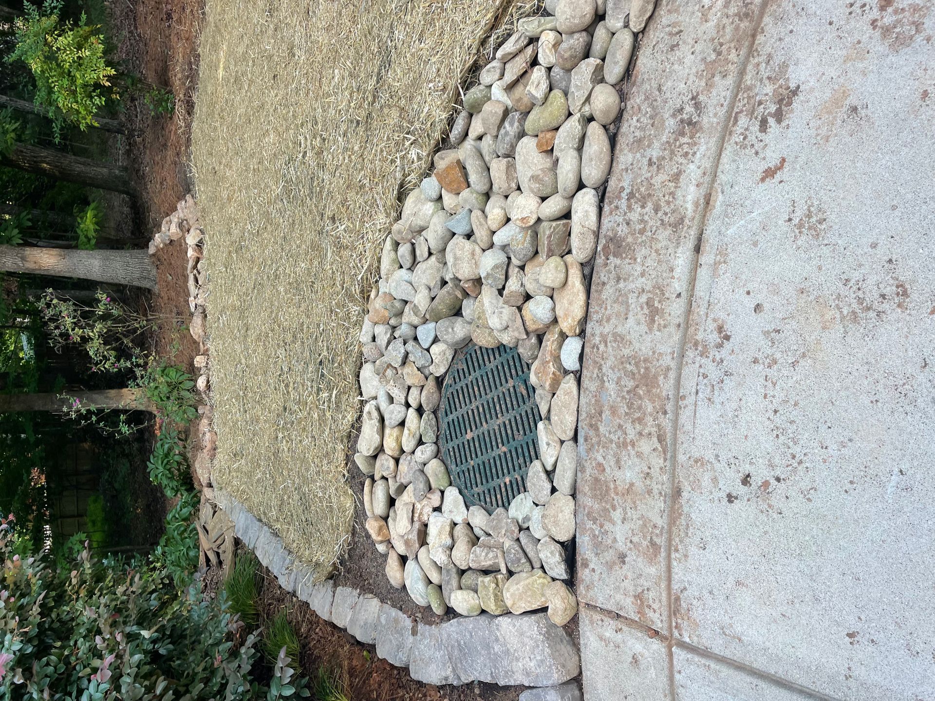 Gravel and stone landscaping borders a concrete patio and a drainage grate.