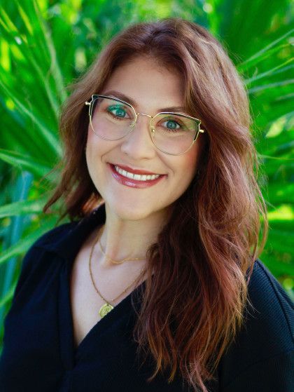 Smiling woman with glasses and long brown hair, standing outdoors with green foliage background