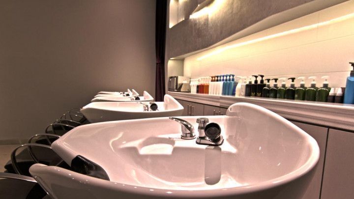 Modern salon wash station with white sinks, black chairs, and shelves of hair products under warm lighting