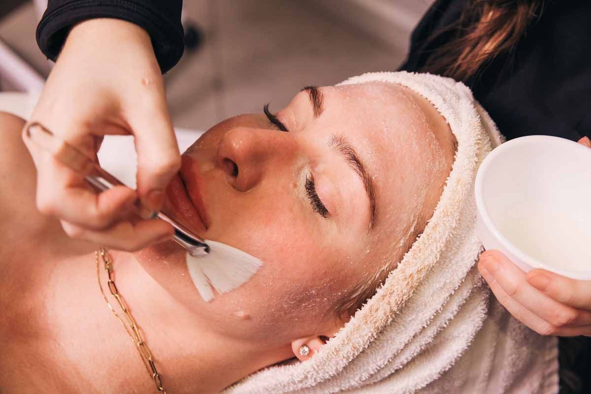 Facial treatment with a cream mask being applied to a client lying down, holding a small bowl and brush