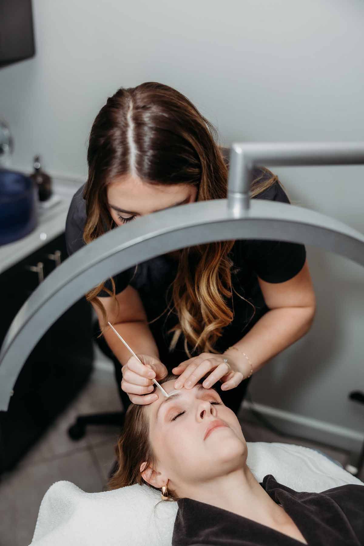 Esthetician performing eyebrow treatment on a client under a circular light in a salon