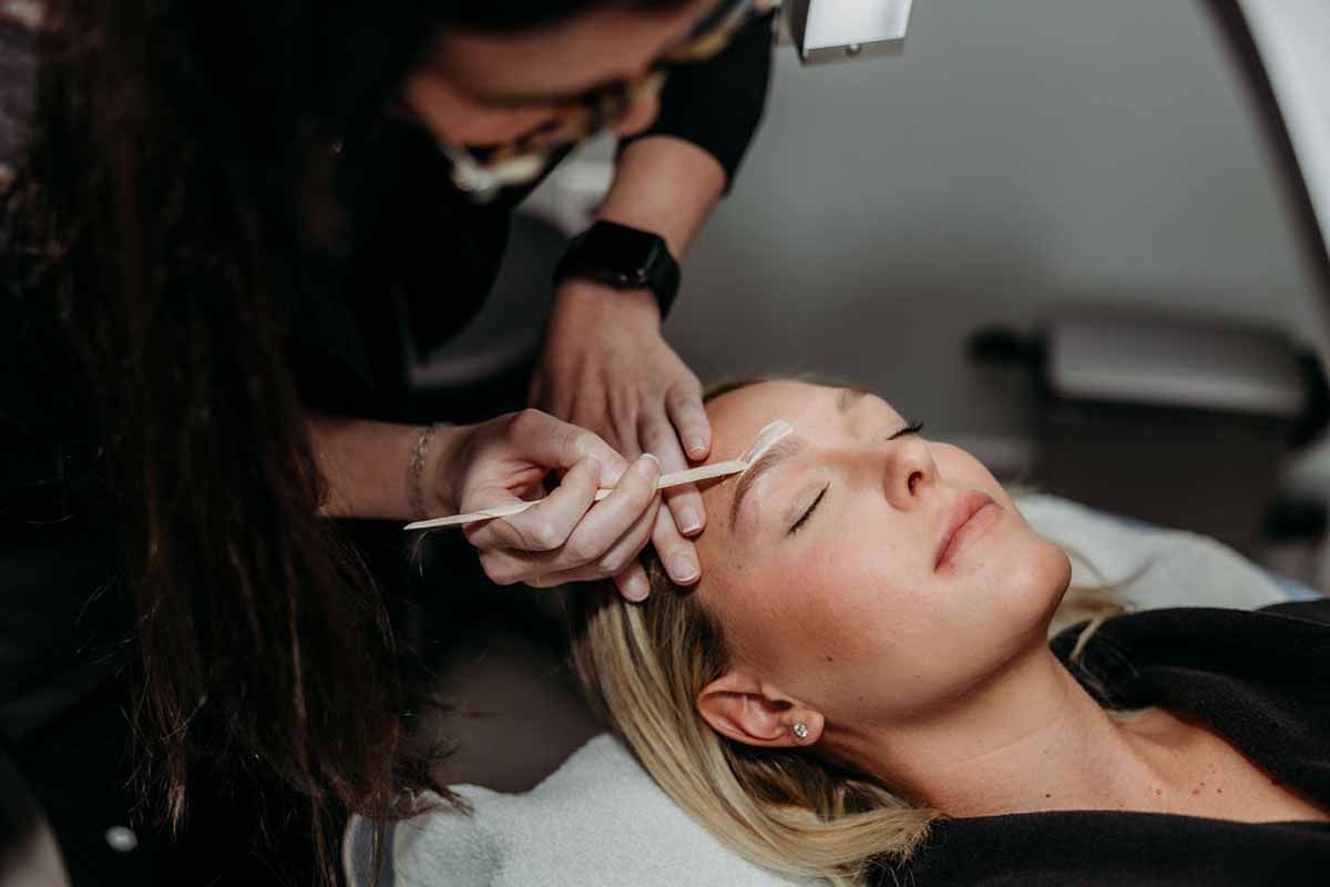 Cosmetologist applying eyebrow treatment to a reclining client in a salon