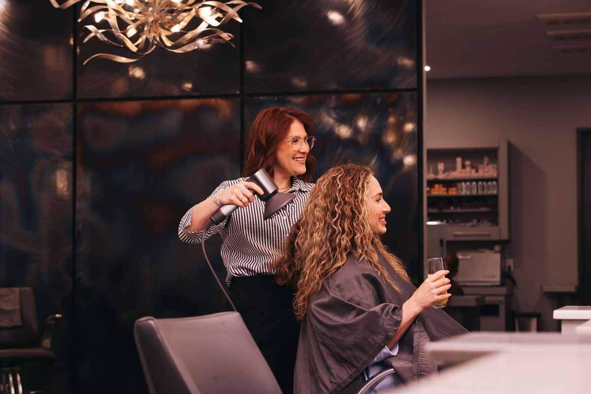 Woman styling another woman's hair in a salon, with shelves and warm lighting in the background.