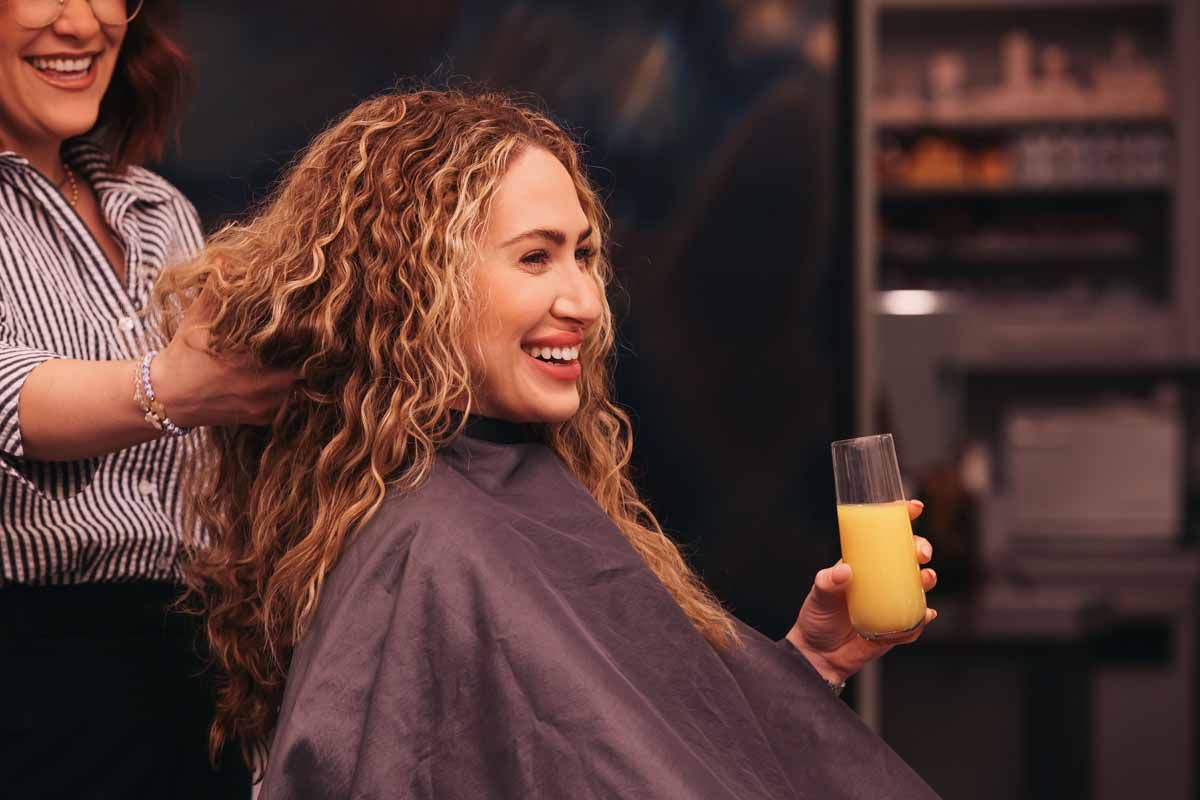 Woman smiling during a salon haircut, holding a yellow drink.