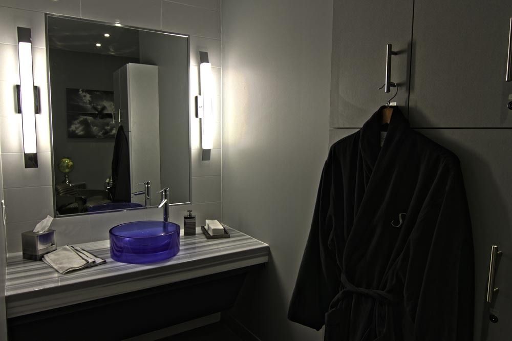Dim bathroom with a lit mirror, sink, purple bowl, and a dark robe hanging nearby.