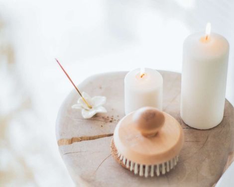 Spa items on a wooden tray with lit candles, incense, and a wooden brush in a soft, bright setting