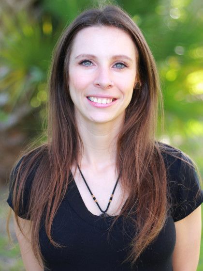 Smiling woman with long brown hair in a black top, standing outdoors against a blurred green background