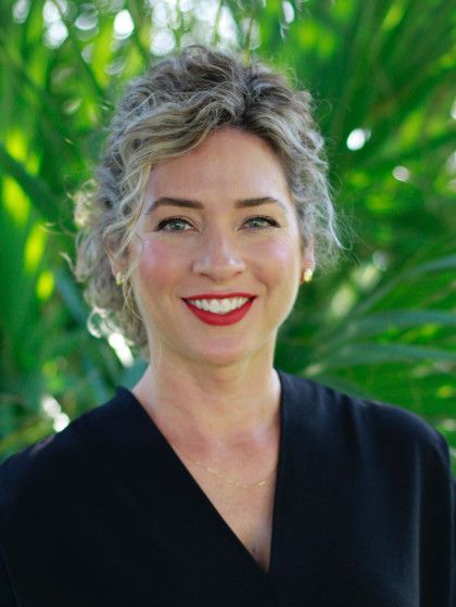 Portrait of a smiling woman with curly gray hair in a black top, outdoors with green foliage background