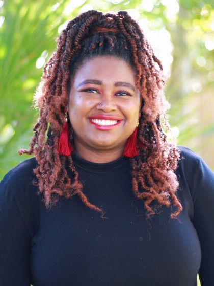 Smiling person in a black top with red earrings, outdoors with green foliage background