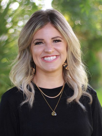 Smiling woman with blonde hair wearing a black top and layered necklaces outdoors with blurred greenery background