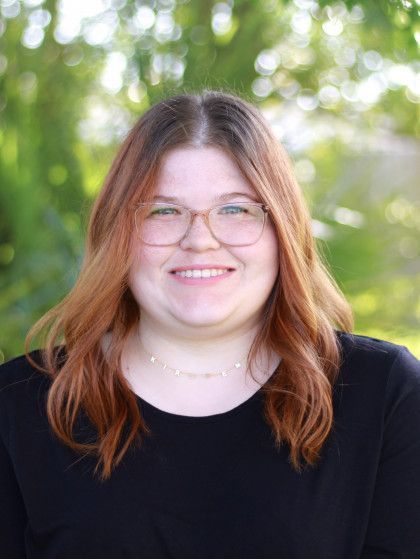 Smiling person with glasses and long reddish hair in a black top, standing outdoors against a leafy green background