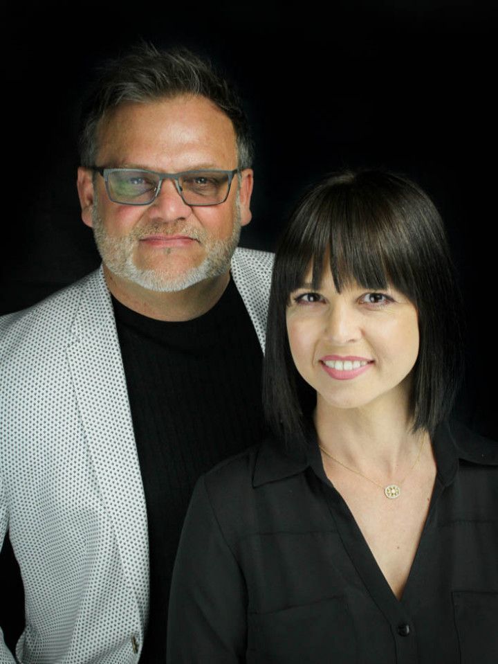 Two people posed against a black background, smiling, one in glasses and a light blazer, one in a black blouse.