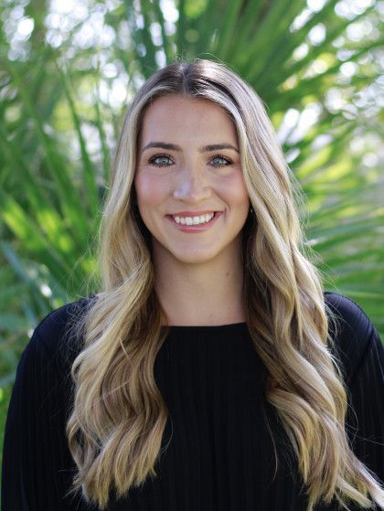 Smiling woman with long blonde hair in a black top, standing outdoors in front of green palm leaves