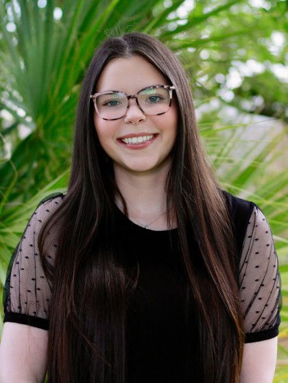 Smiling woman with long dark hair and glasses, standing outdoors in front of green foliage.