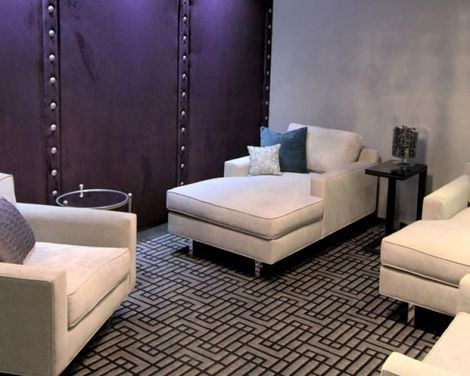 Modern lounge with beige armchairs and ottoman on a patterned rug beside dark paneled walls.