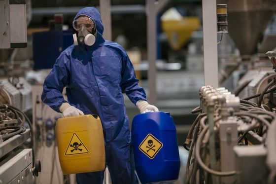 Un uomo con una tuta protettiva tiene in mano due barili di prodotti chimici in una fabbrica.
