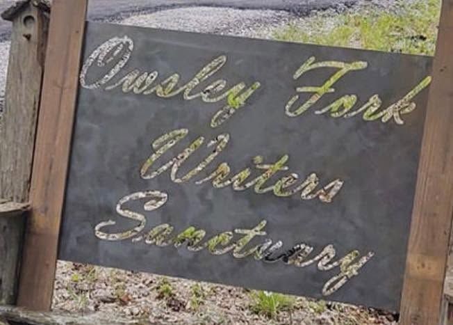 Wooden sign reading “Owsley Fork Writers Sanctuary” in script lettering on a stone wall