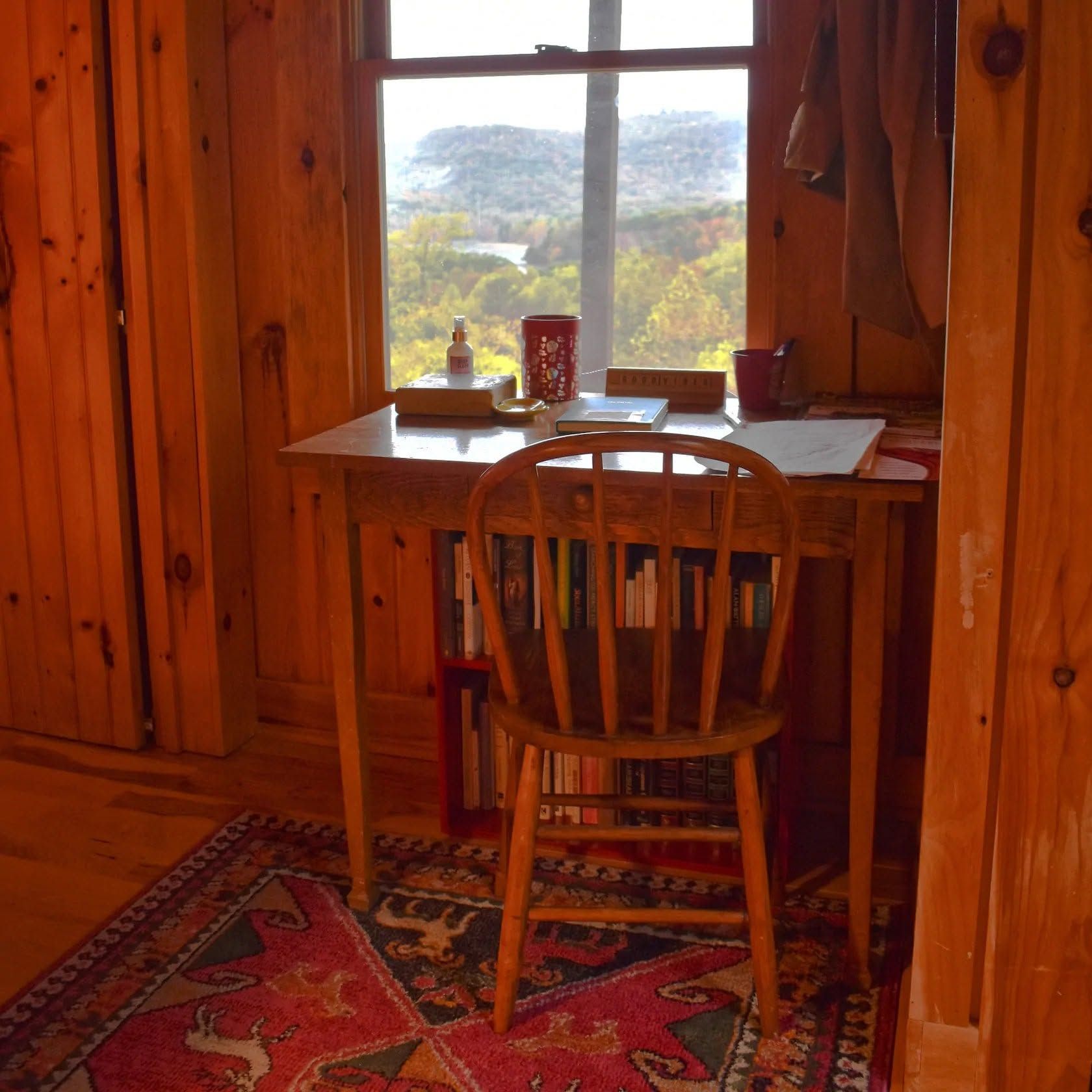 Wooden desk and chair by a window in a cozy cabin, with a view of trees outside.