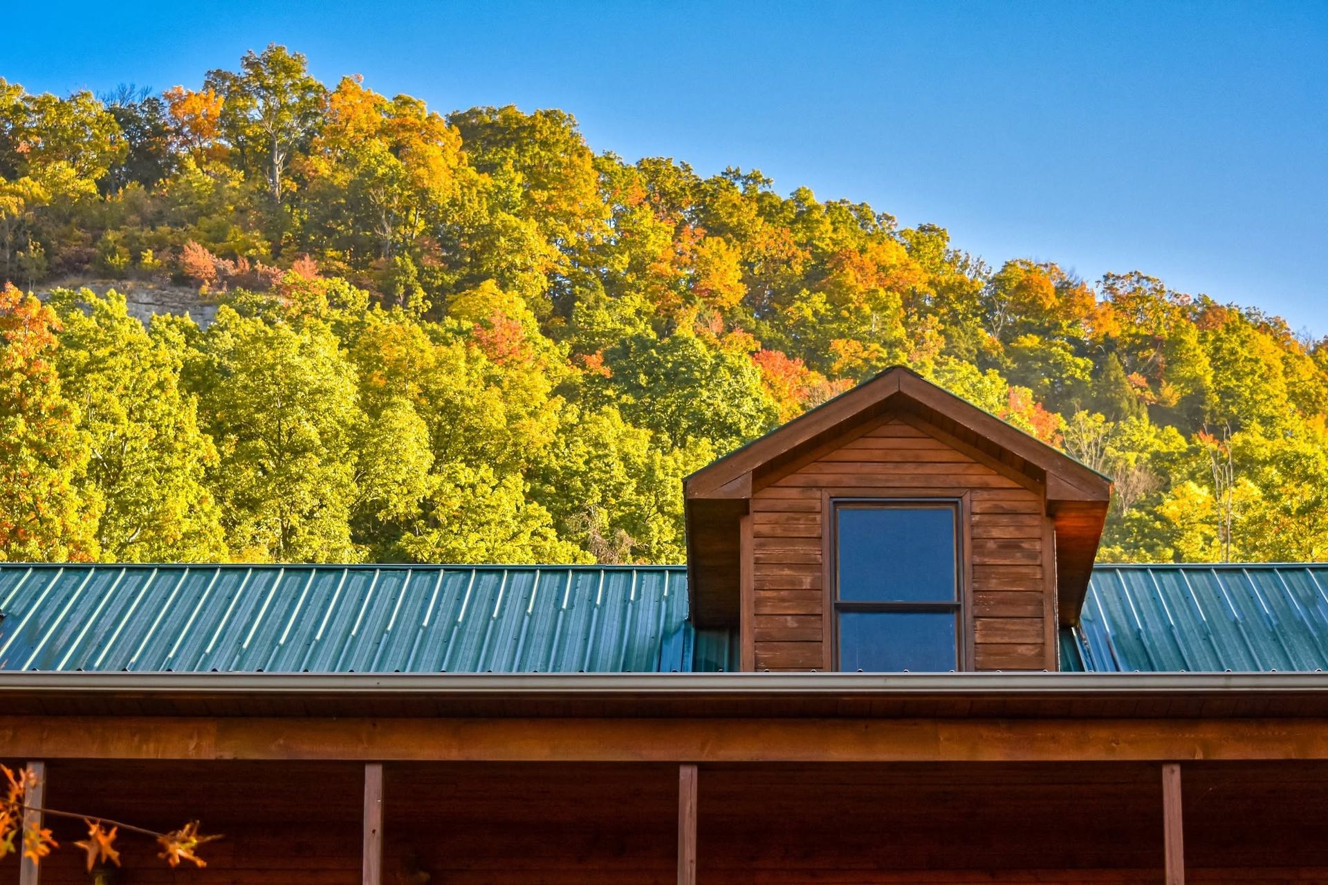 Rustic cabin roof with a dormer in front of a sunlit autumn hillside and blue sky