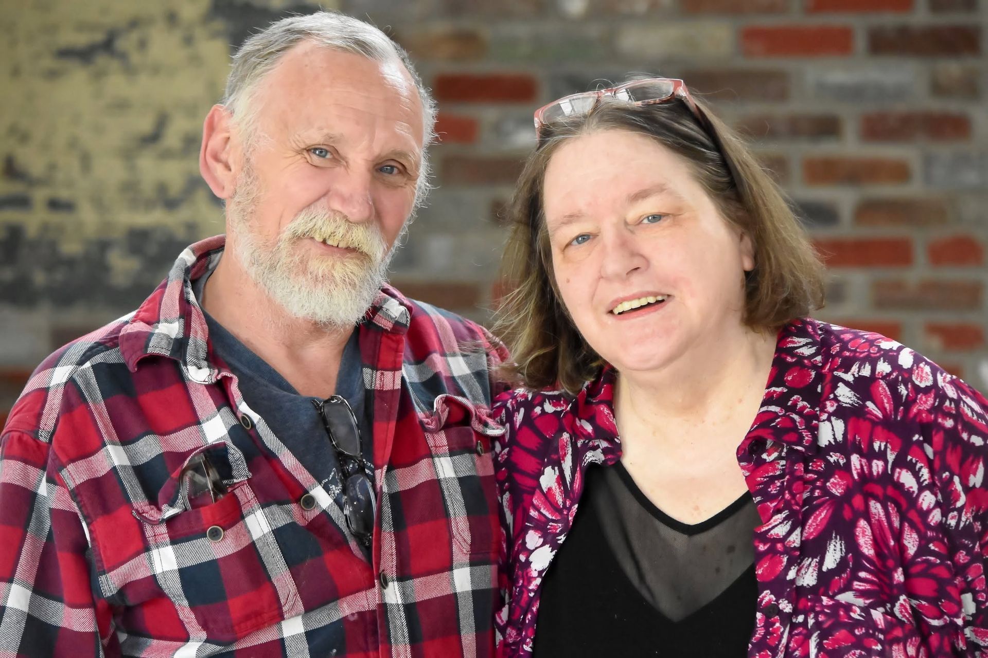 Two people smiling and standing close together in front of a brick wall, wearing plaid and floral tops.