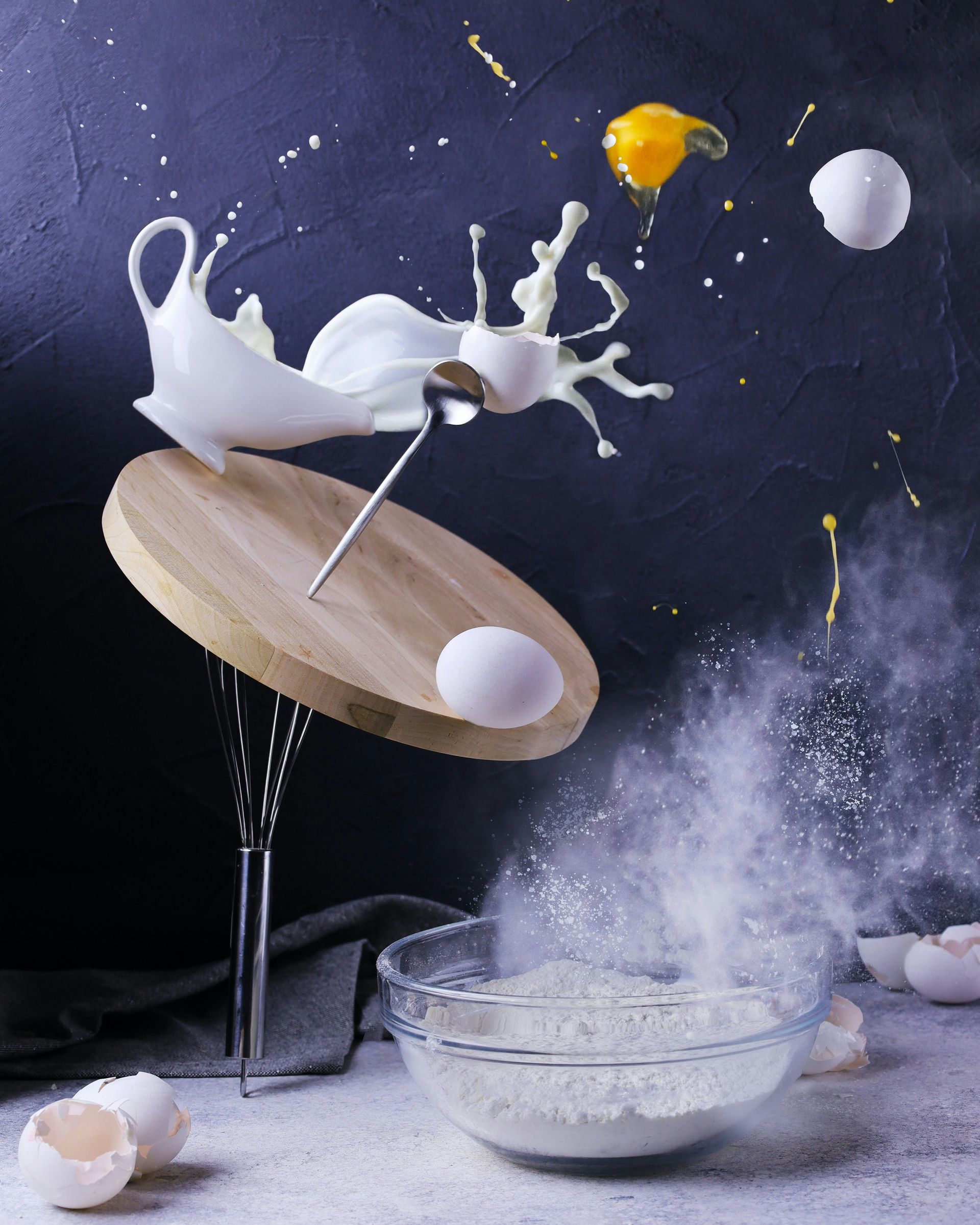Milk splashing from a tipped bowl, with flour dust and broken eggs against a dark background