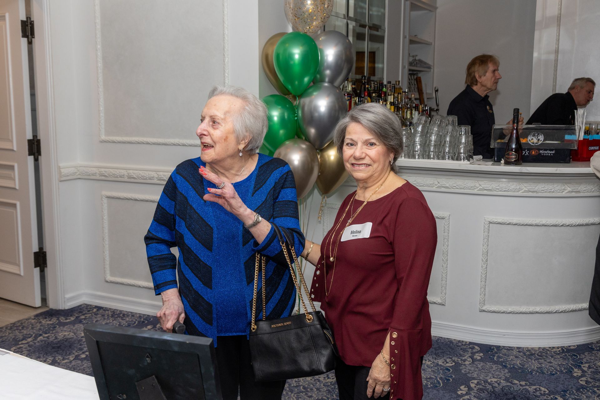 Two women at a party, one in blue, one in maroon, near a bar with balloons.