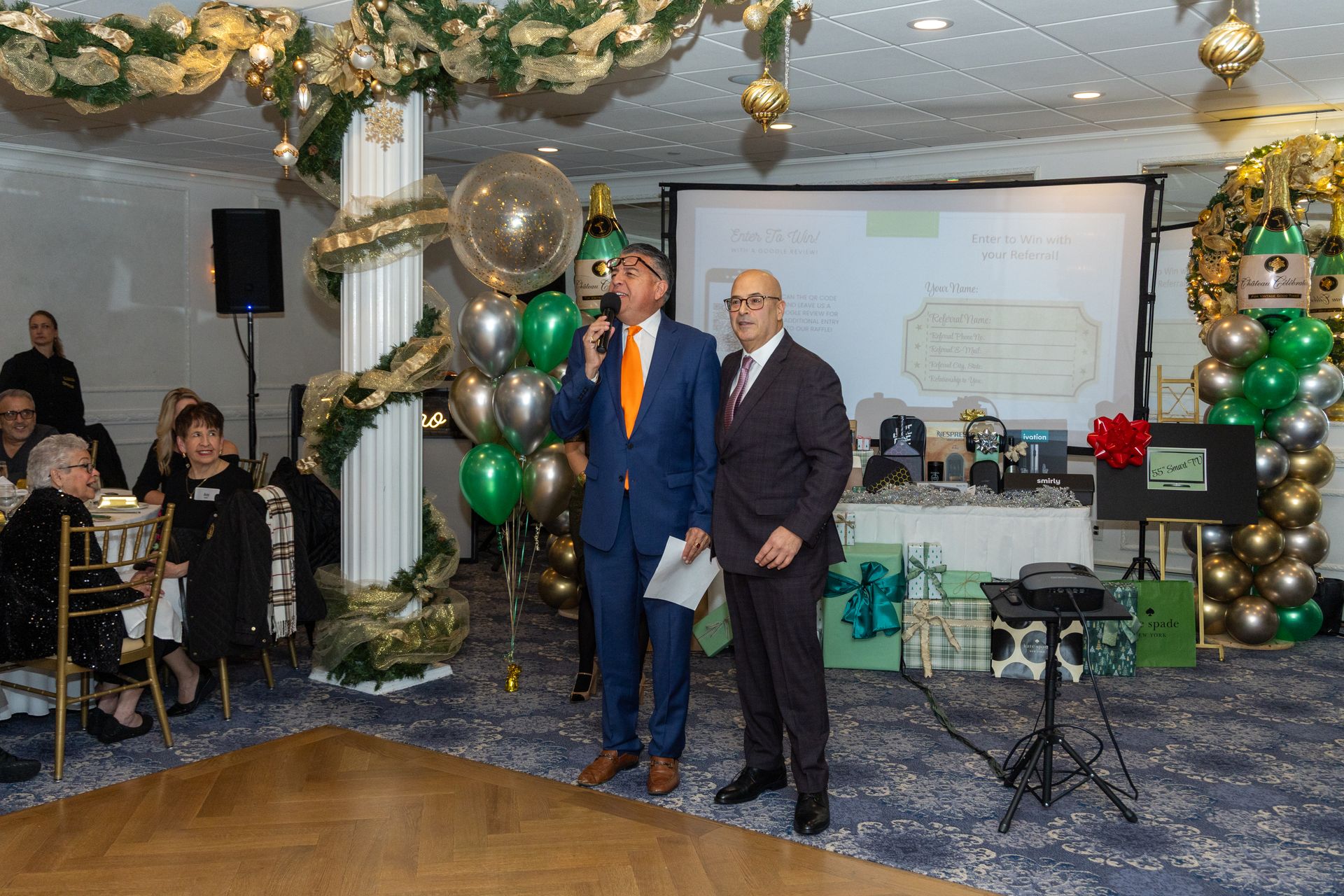 Two men at podium, speaking at event. Room decorated with balloons, greenery, and presents.