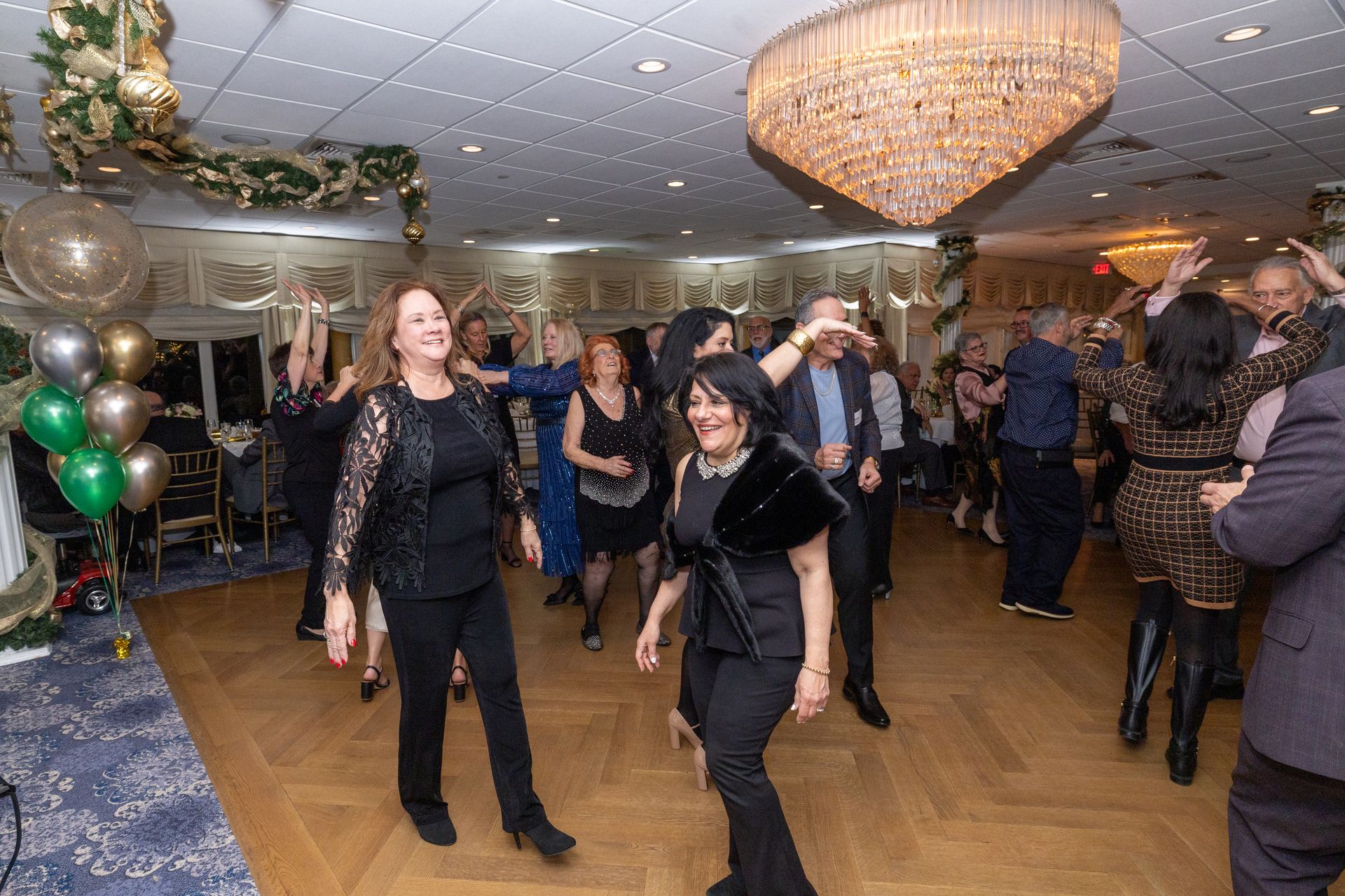 People dancing in a decorated ballroom with a chandelier, balloons, and garland.