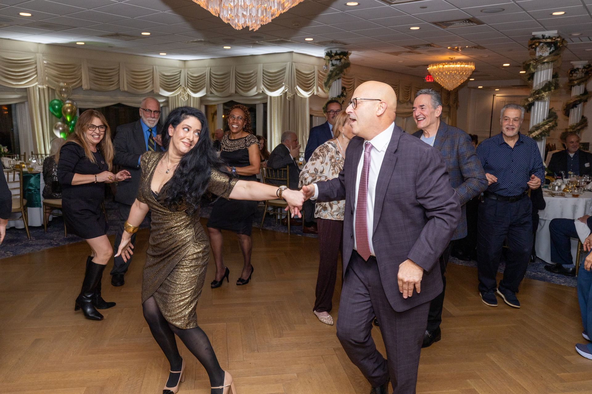 People dancing at a party; gold and black dresses, suits, chandelier, decorated room.