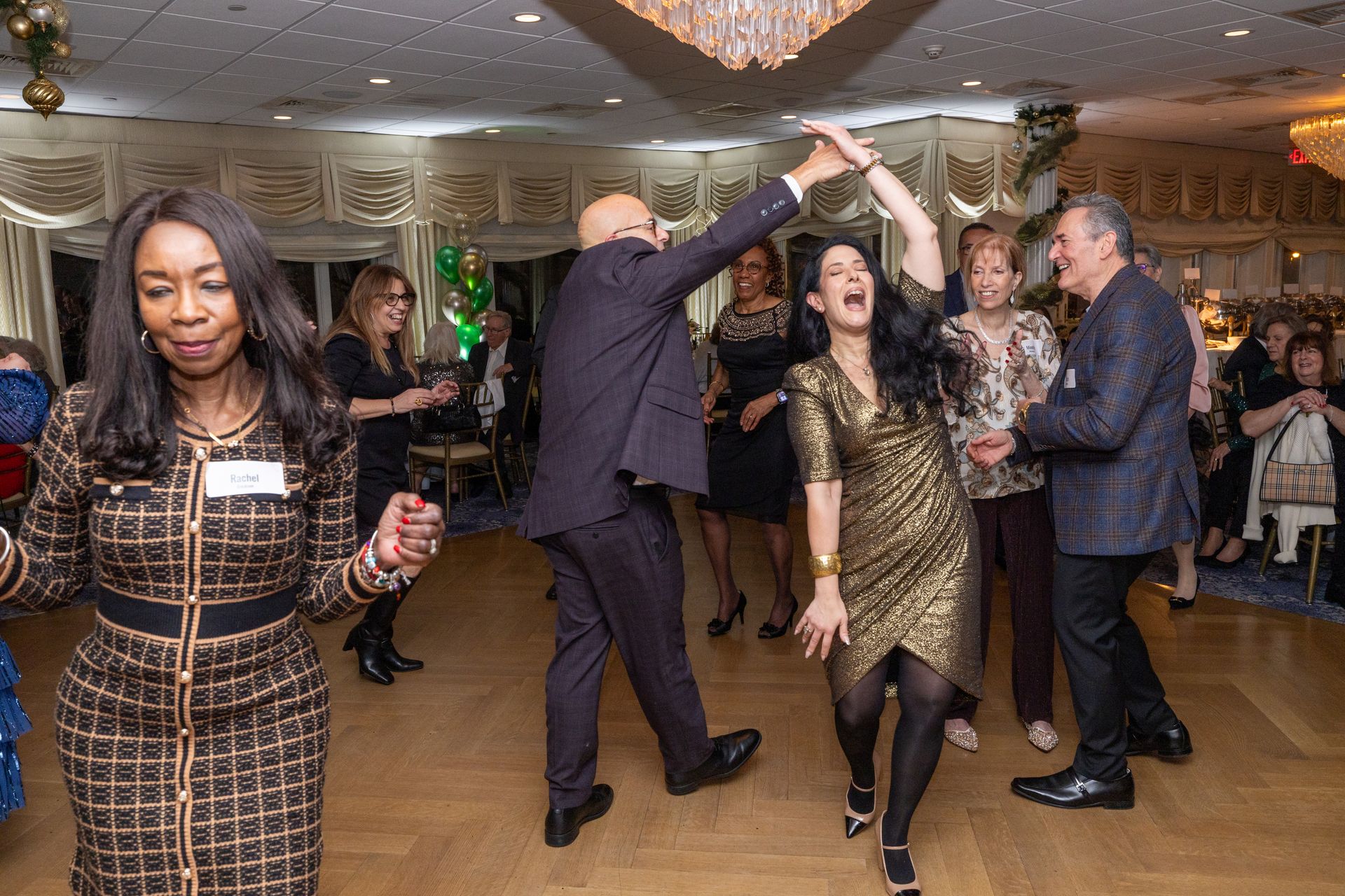 People dancing in a ballroom; woman in gold dress laughs, man raises her hand, others watch.