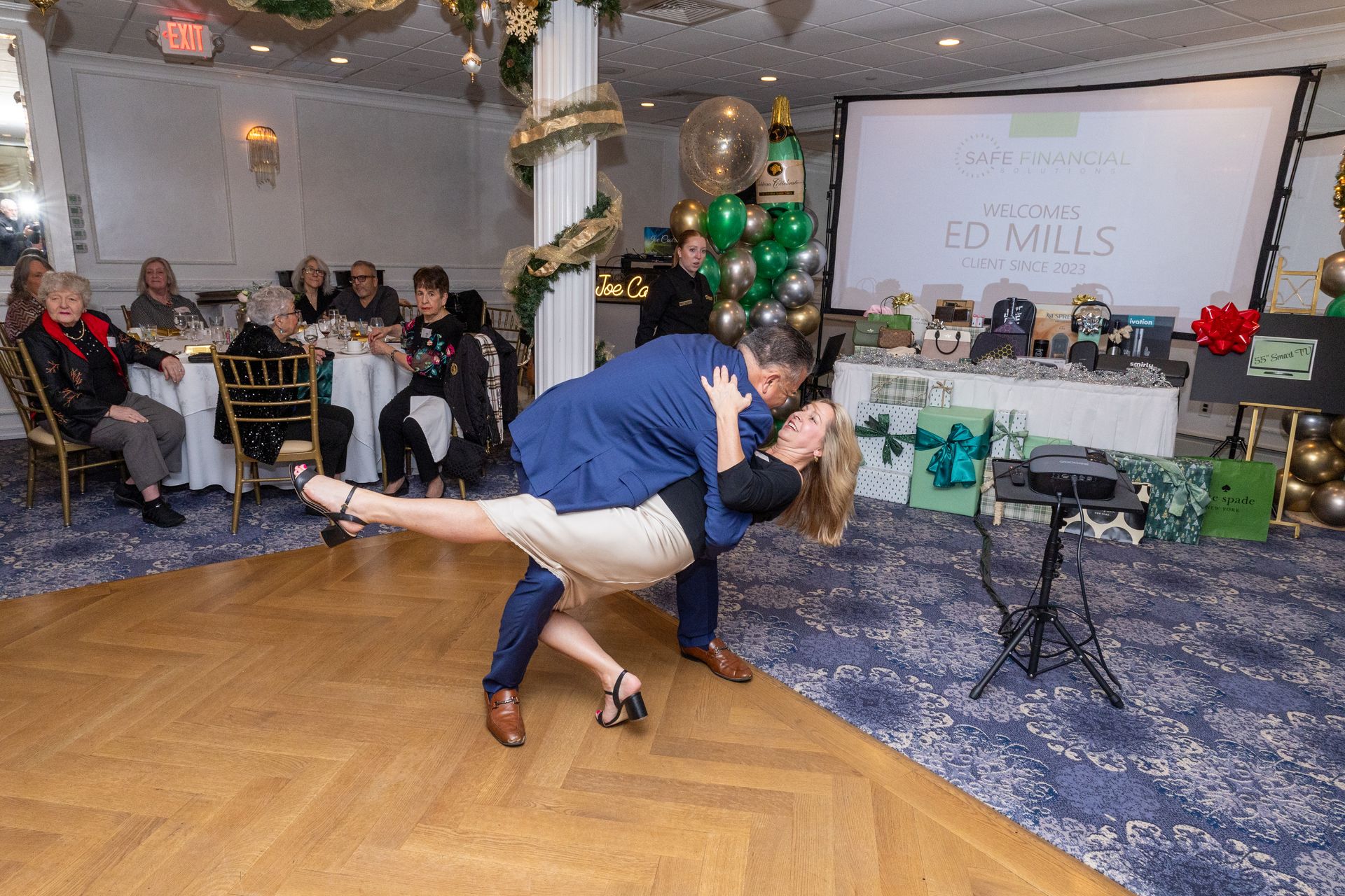 Couple dancing at an event, man lifting woman. People seated at tables, decorated room, projection screen in background.