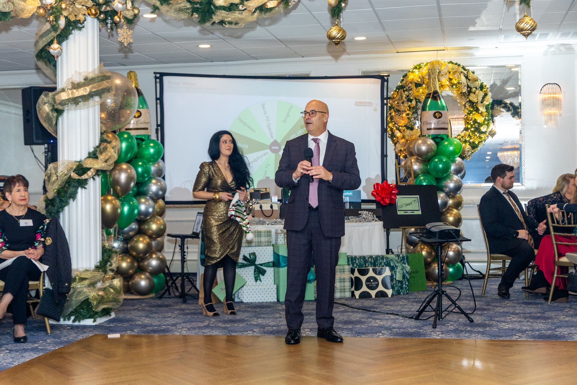 People on stage at an event, speaking. Green and gold decorations. A presentation screen is in the background.