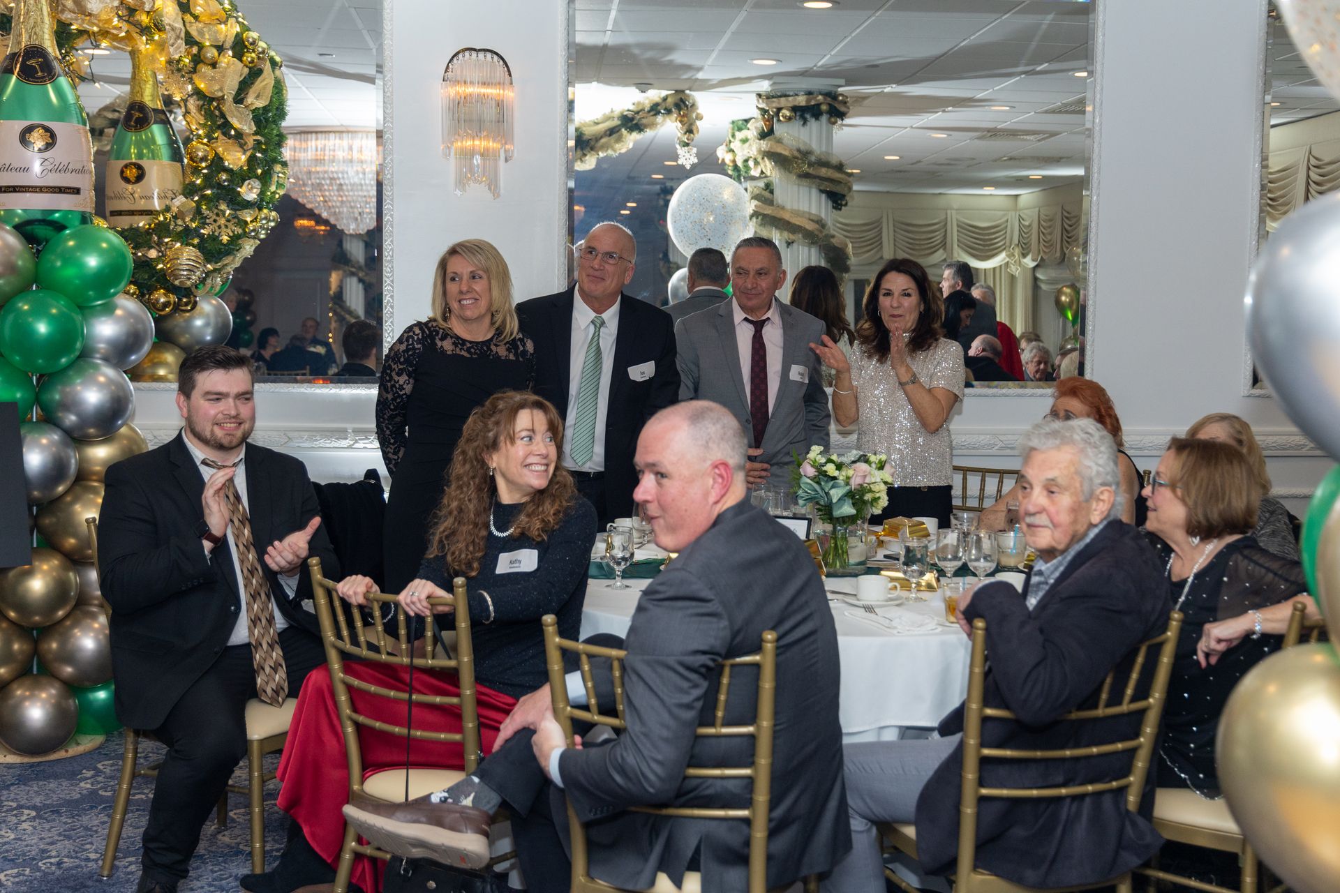 People at a celebratory event, seated at tables and standing. Balloons and festive decor are in the background.