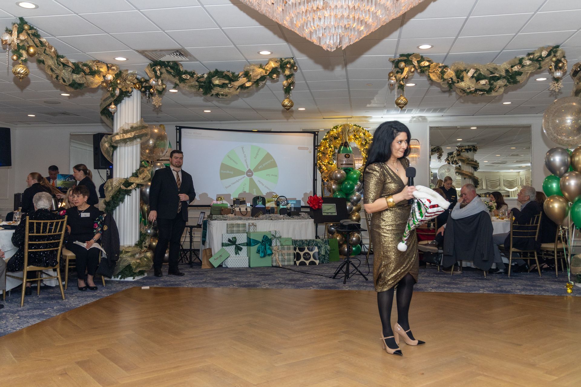 Woman in gold dress holds gift, on dance floor. Festive decorations and event attendees in background.