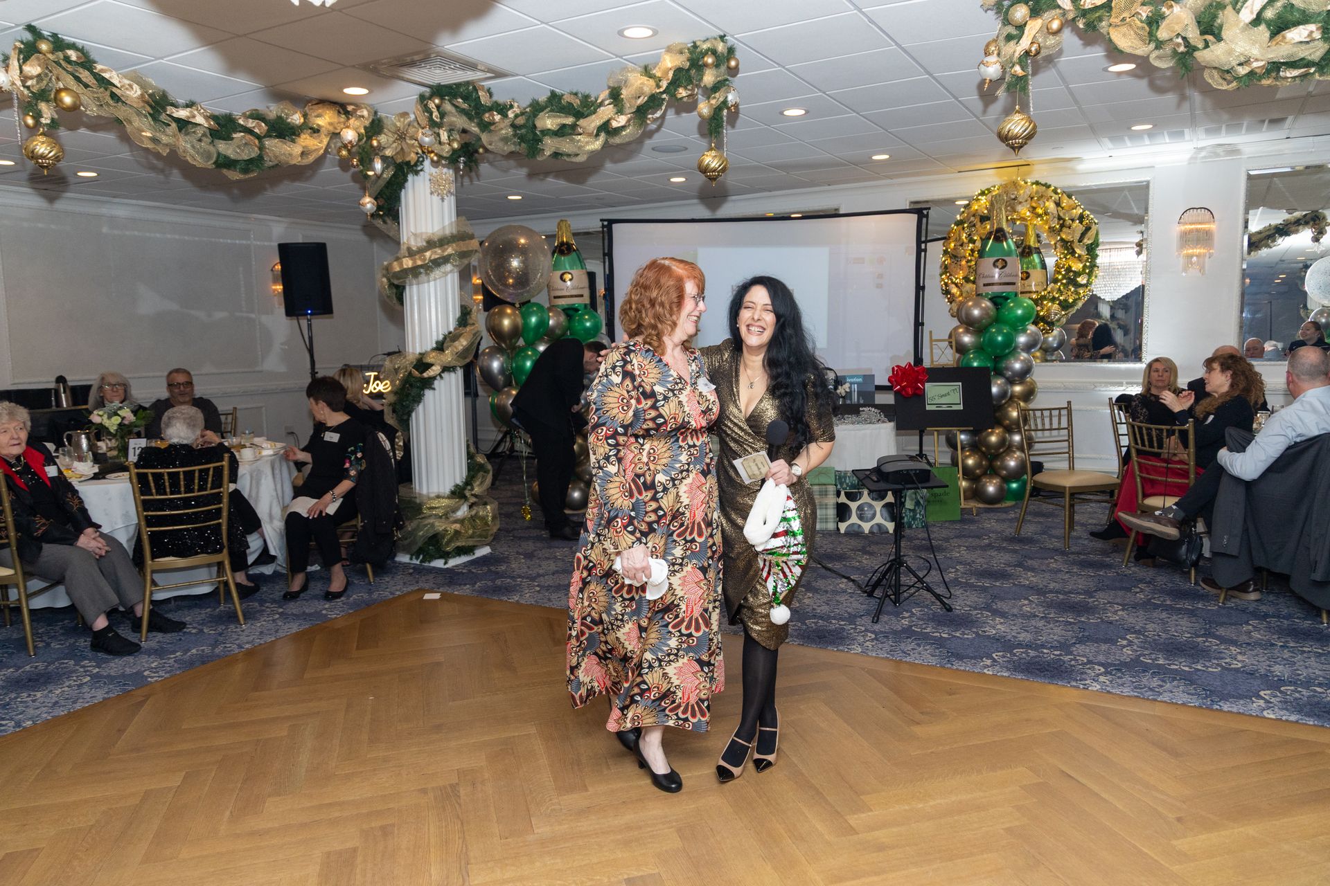 Two women embrace on a dance floor, decorated for an event. One holds a small gift.