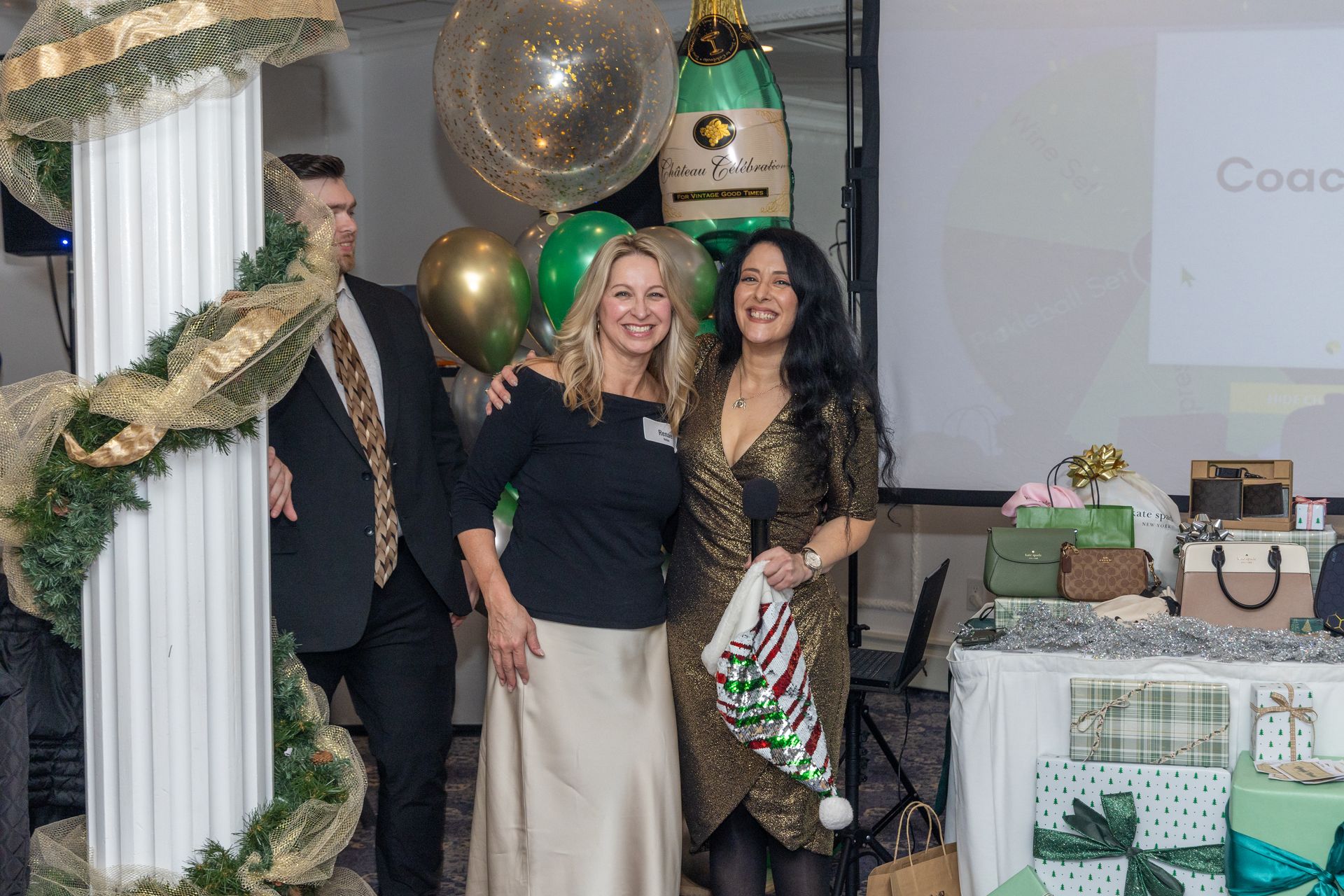 Two women smiling, posing at event. Christmas decor in background, gifts on table.