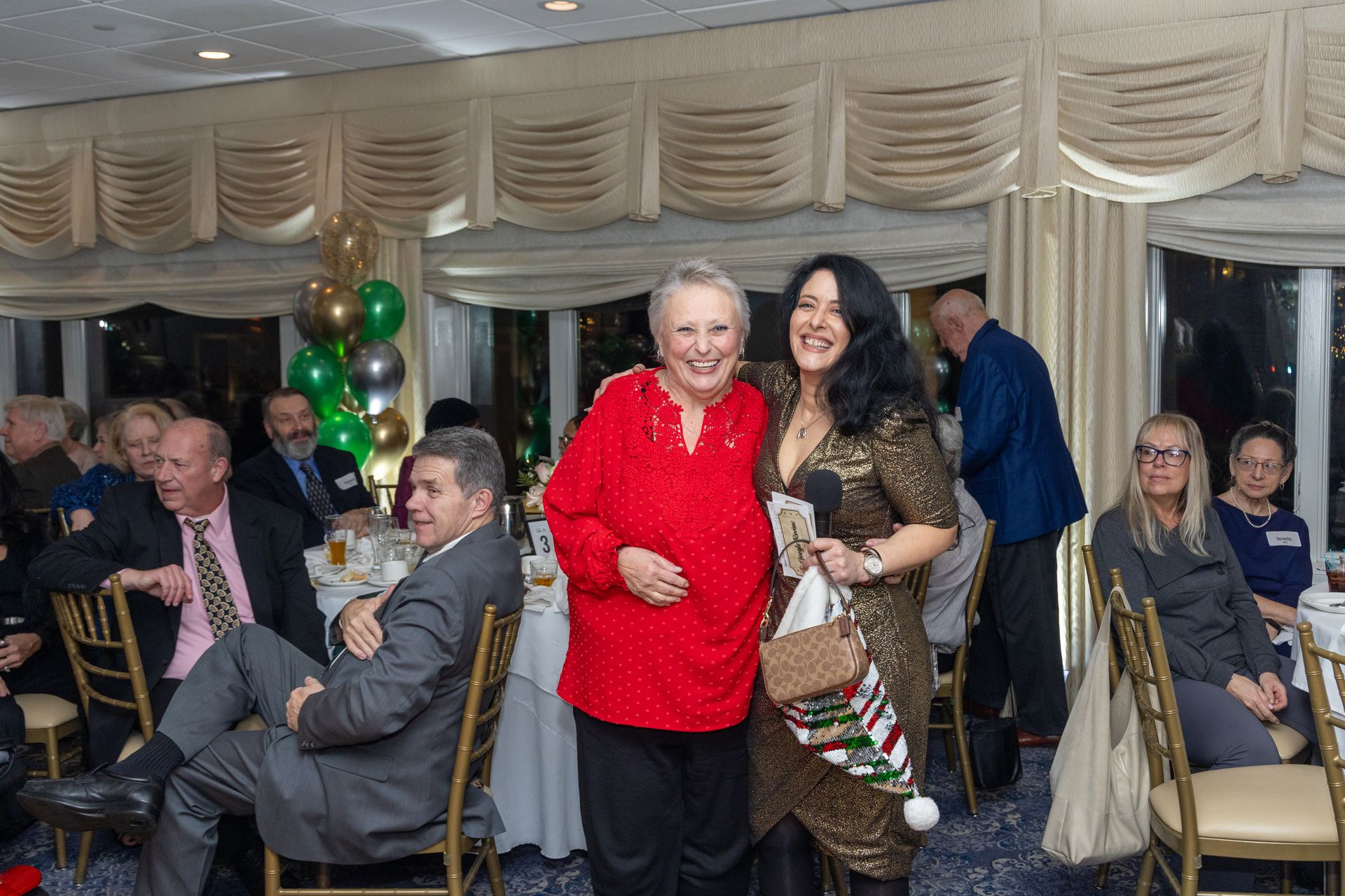 Two women smiling at a Christmas party. One wears red, the other gold, holding a stocking. Others seated at tables.