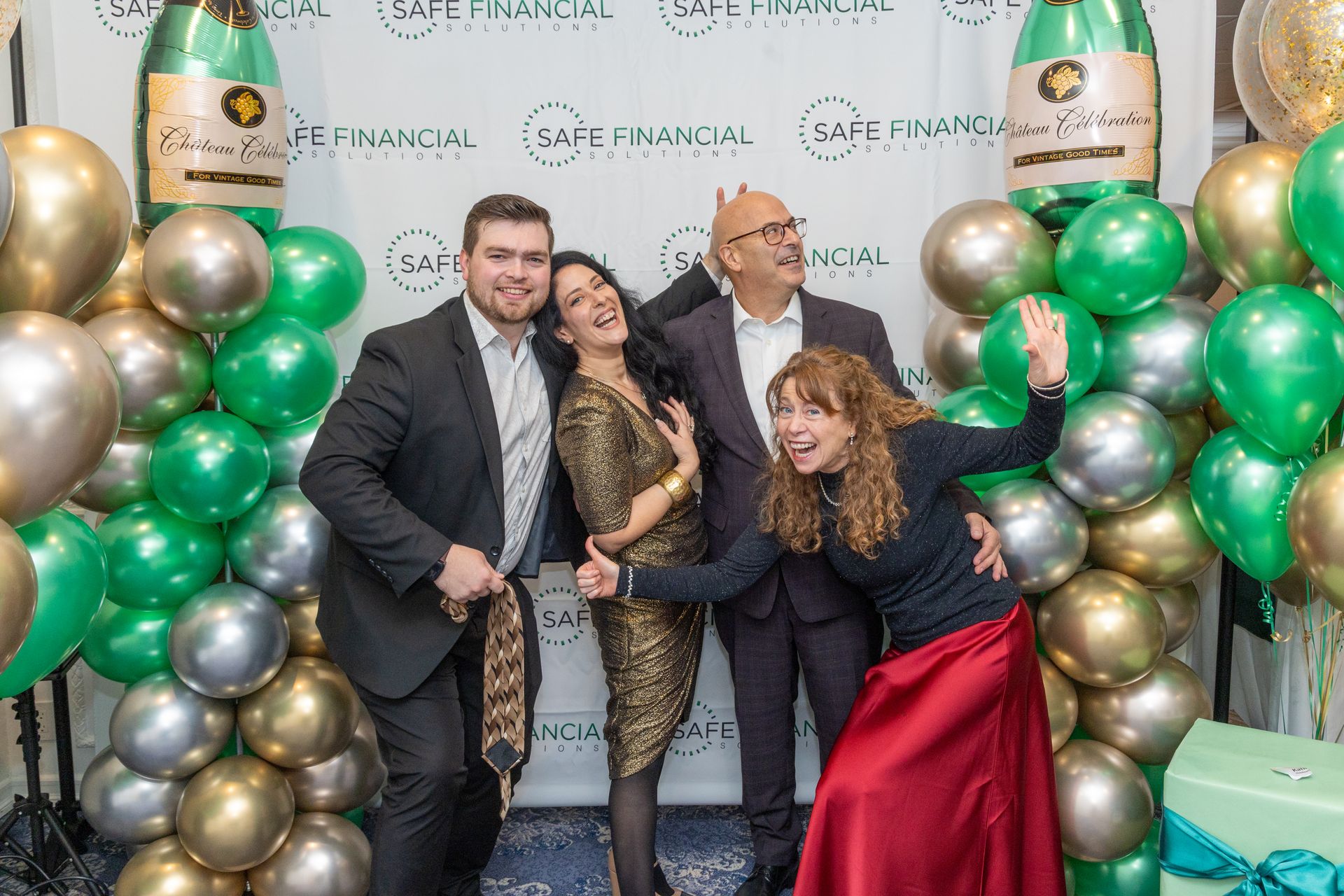 Four people pose in front of a backdrop with balloons. Green, gold, and silver balloons decorate the area.