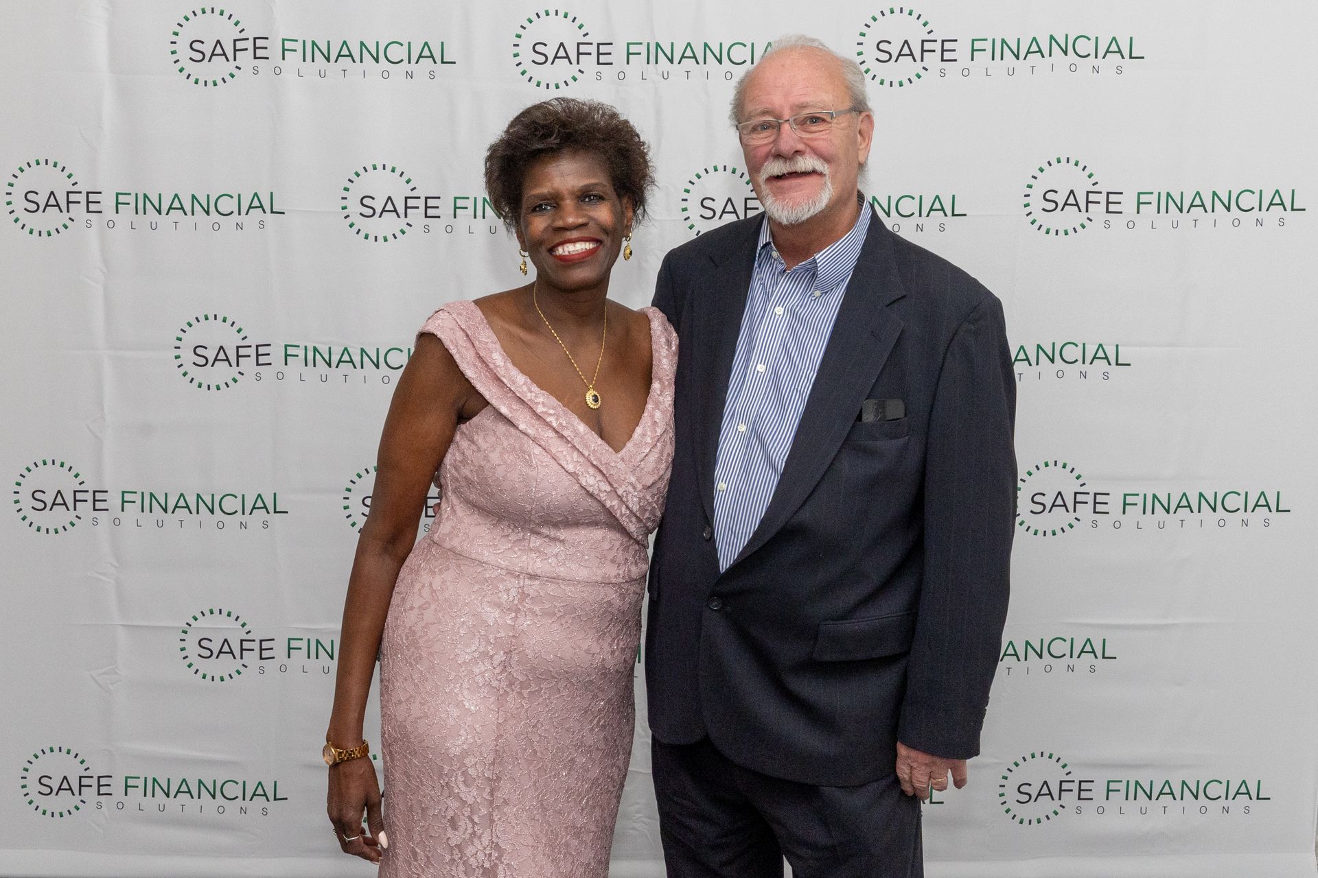 A woman in a pink dress and a man in a blazer stand in front of a backdrop with the words 