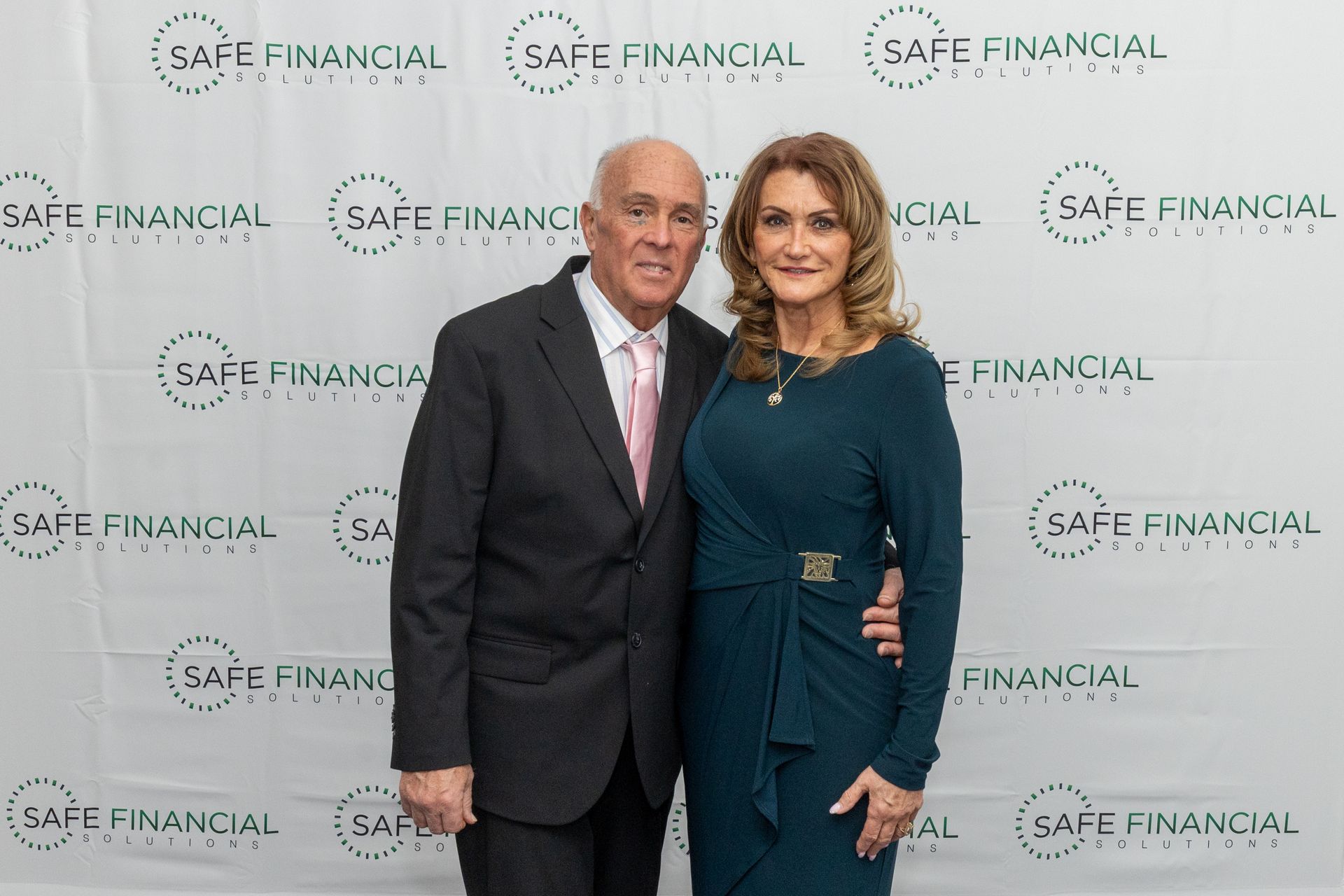 A man and woman pose in front of a backdrop with a company logo. The woman wears a teal dress, and the man wears a suit.