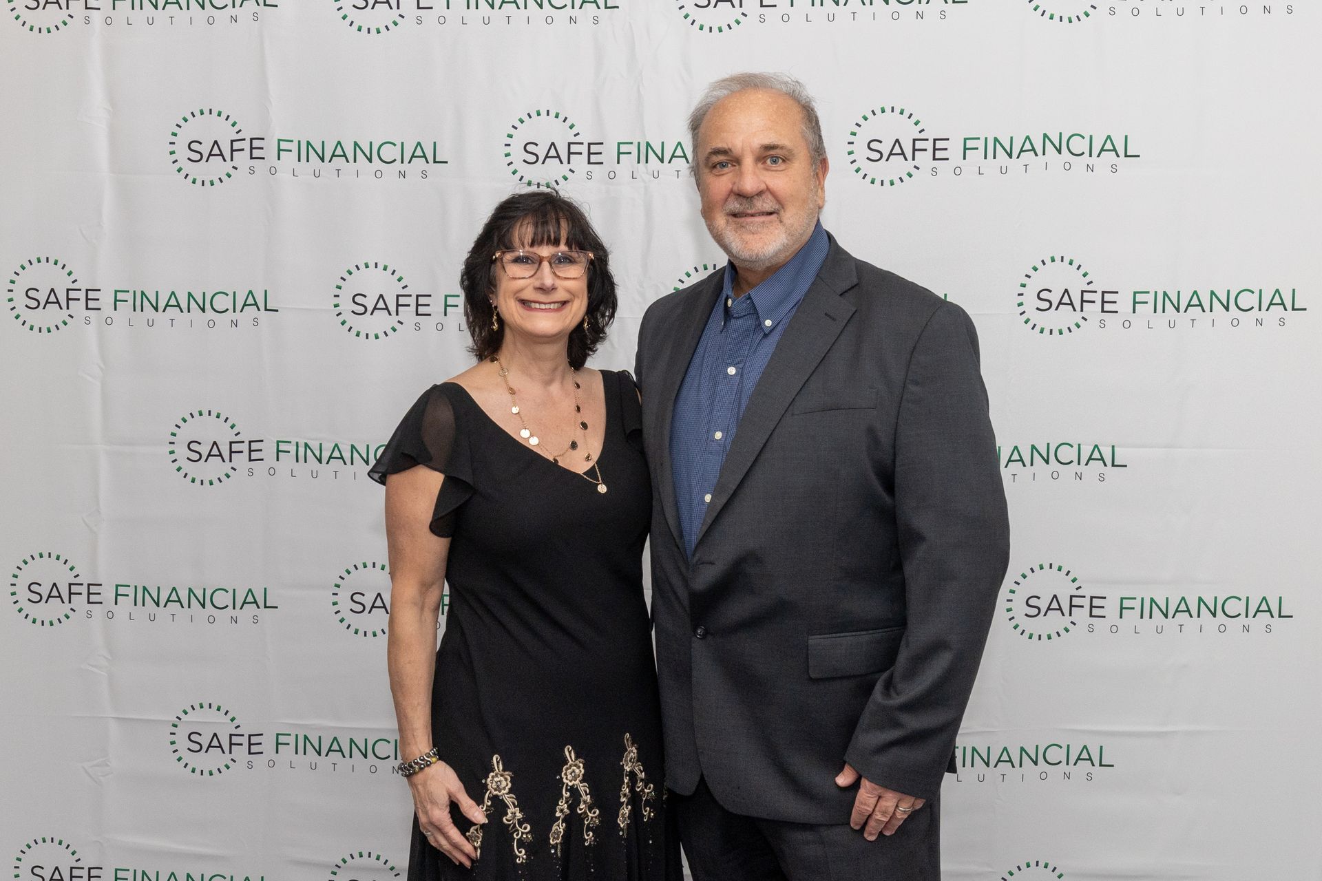 A couple poses in front of a backdrop with logos. The woman wears a black dress and the man a suit.