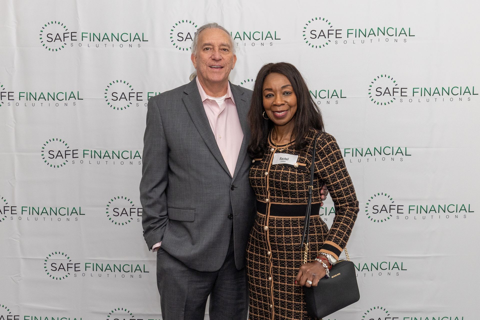 Man and woman pose in front of a backdrop with a company logo. The woman wears a patterned dress and holds a purse.