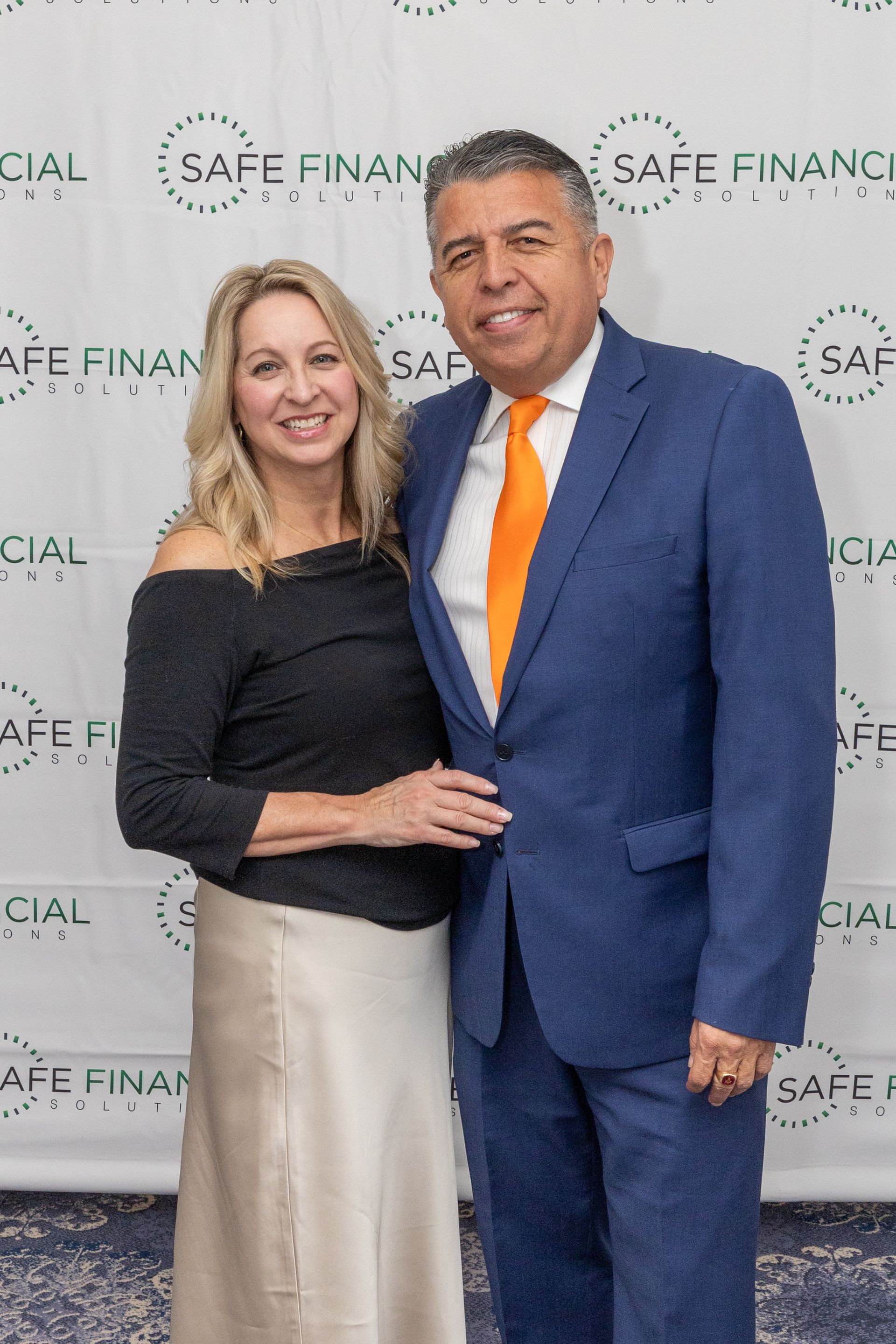 The man wears a blue suit and orange tie. The woman wears a black top and cream skirt.