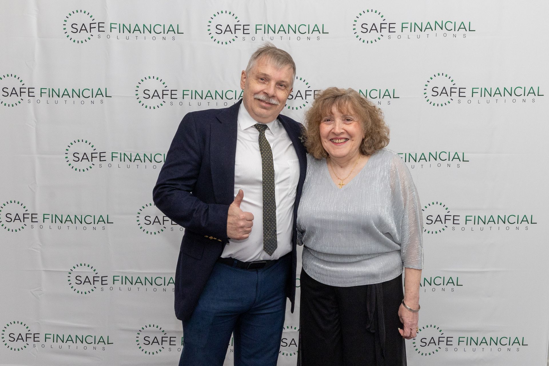 Two people smiling, posing in front of a SAFE FINANCIAL backdrop; man gives thumbs-up.