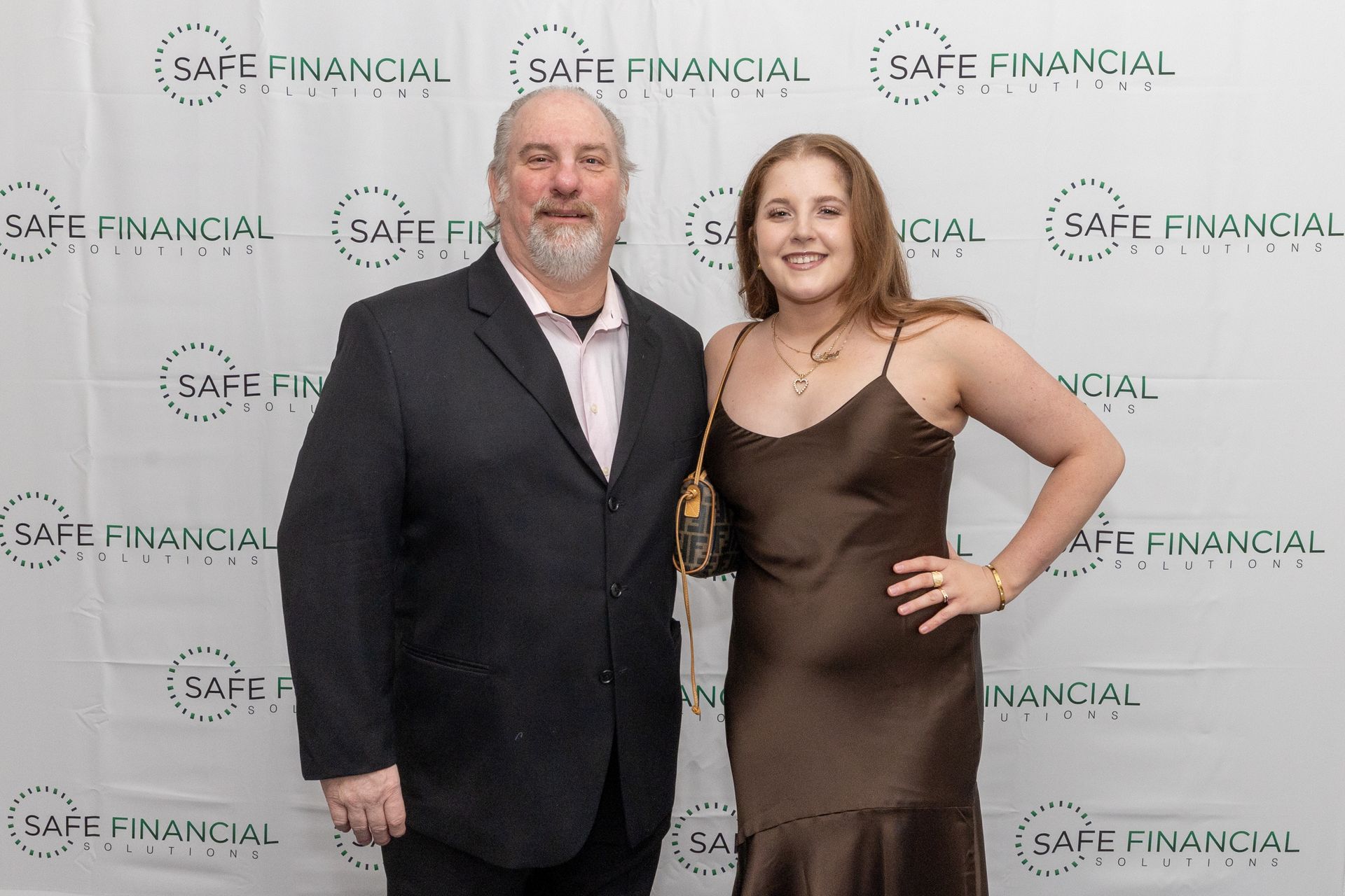 A man in a suit and a woman in a brown dress stand together in front of a backdrop with 