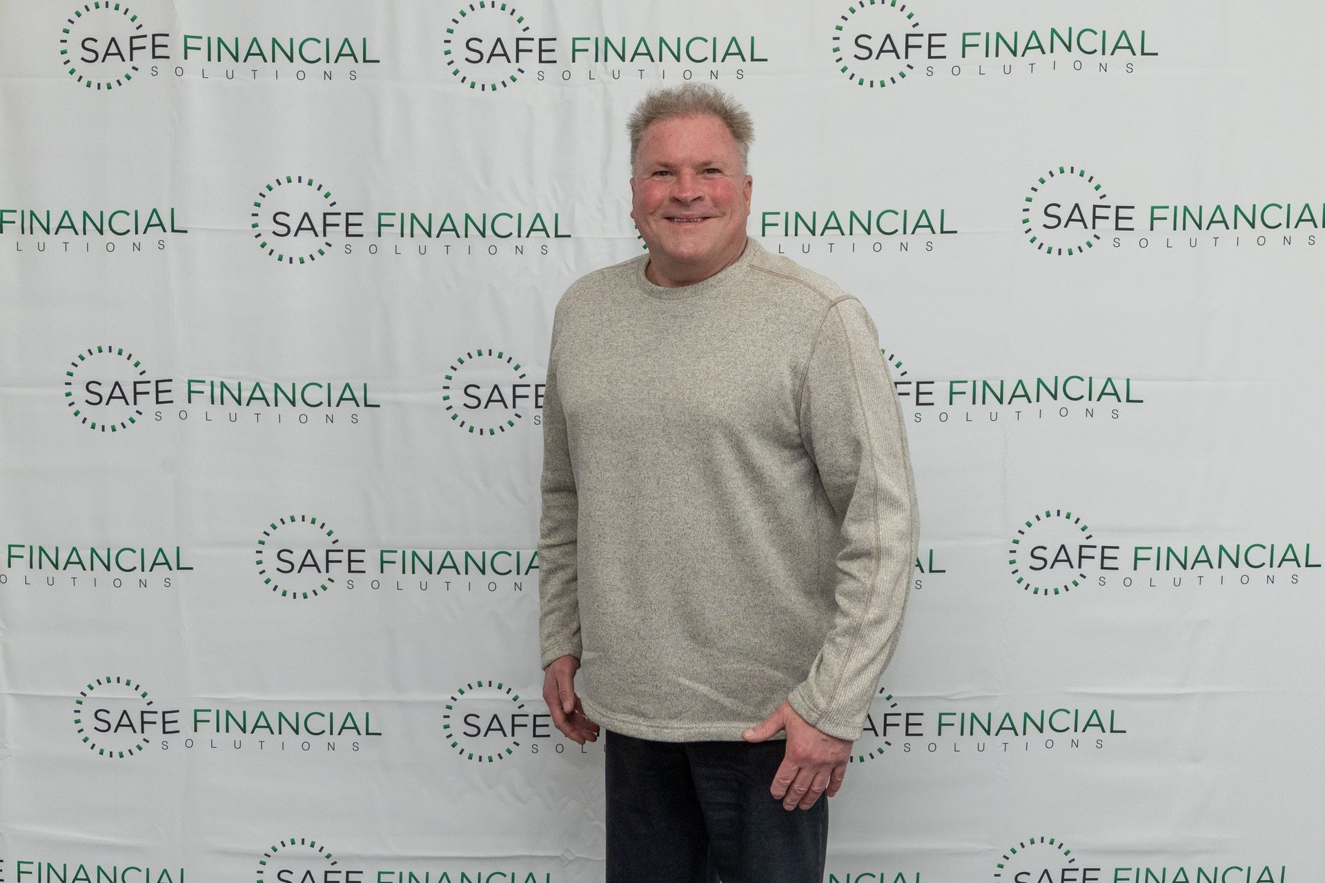 Man in a light-colored sweater smiles in front of a backdrop with 